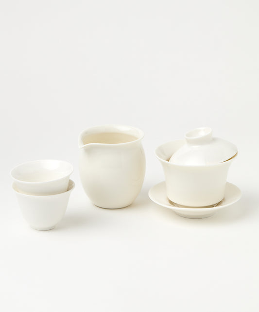 A white porcelain gaiwan with a tilted lid, a pitcher, and two stacked teacups.