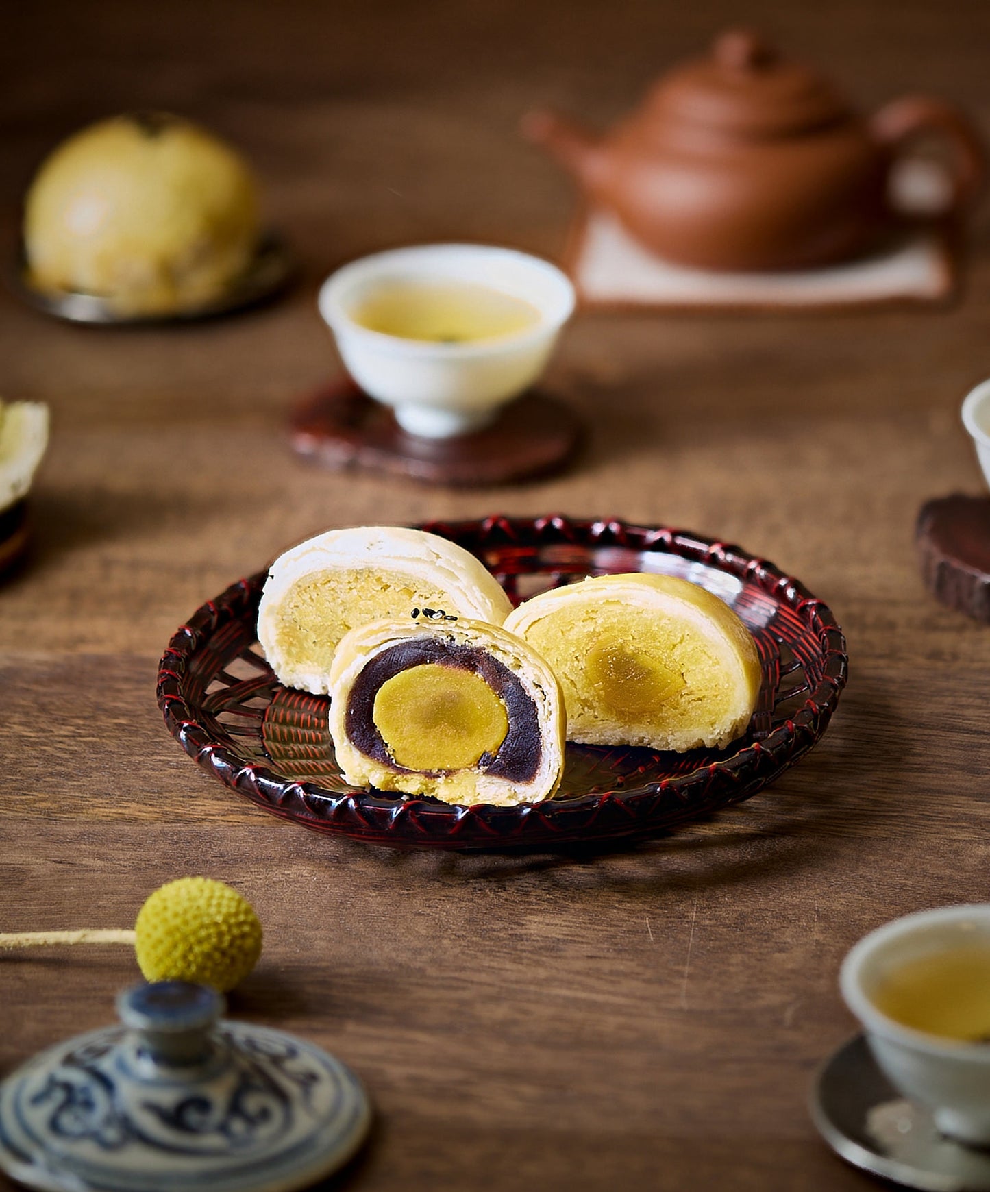 A woven basket holding three mooncake halves with visible fillings, paired with teacups and a teapot on a wooden table.