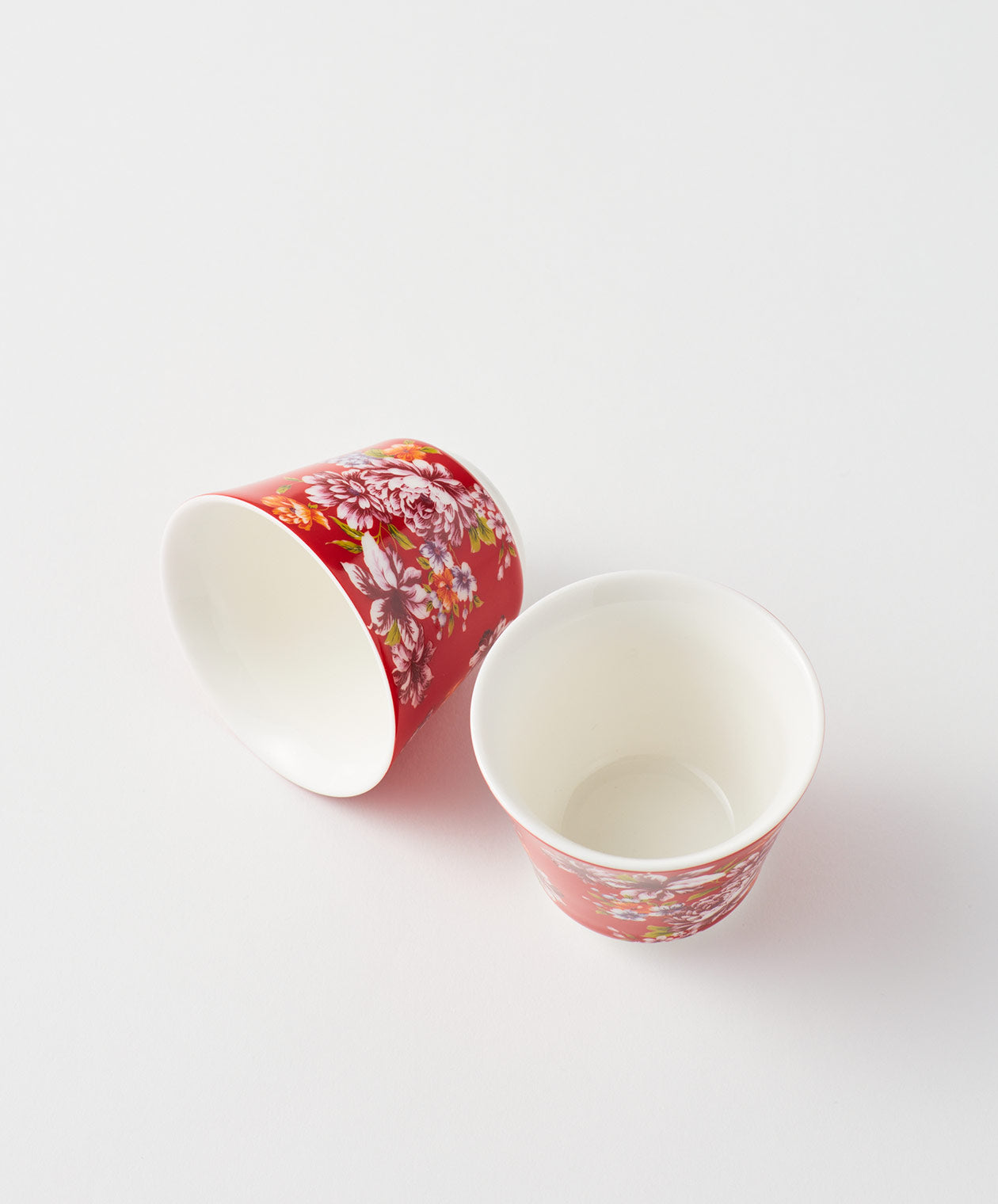 Two red ceramic cups with floral designs, one standing and one tilted.