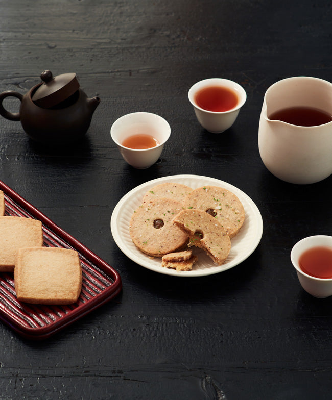 A tea setting with shortbread cookies displayed on a plate and tray, accompanied by teacups filled with tea, a teapot, and a pitcher on a dark wooden table.