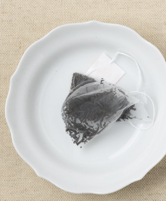 A pyramid-shaped tea sachet filled with dried leaves, displayed on a white plate against a beige background.