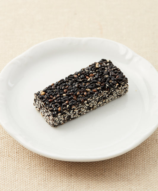 A side view of a black sesame seed bar on a white plate against a beige fabric background.