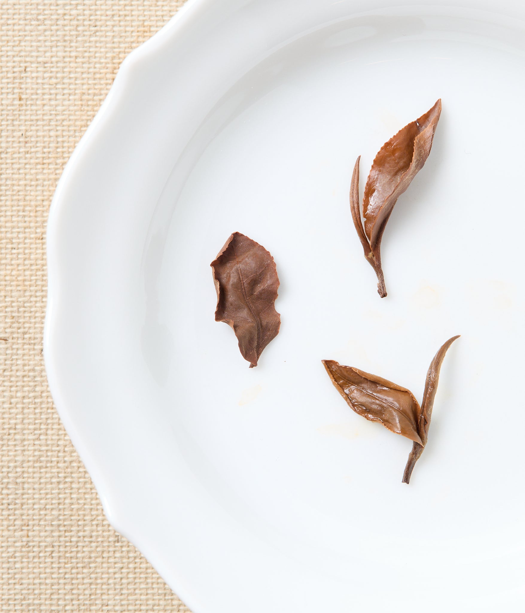 Three tea leaves on a white plate with a beige mat background.