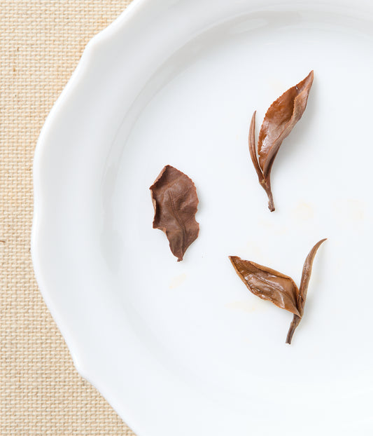 Three tea leaves on a white plate with a beige mat background.