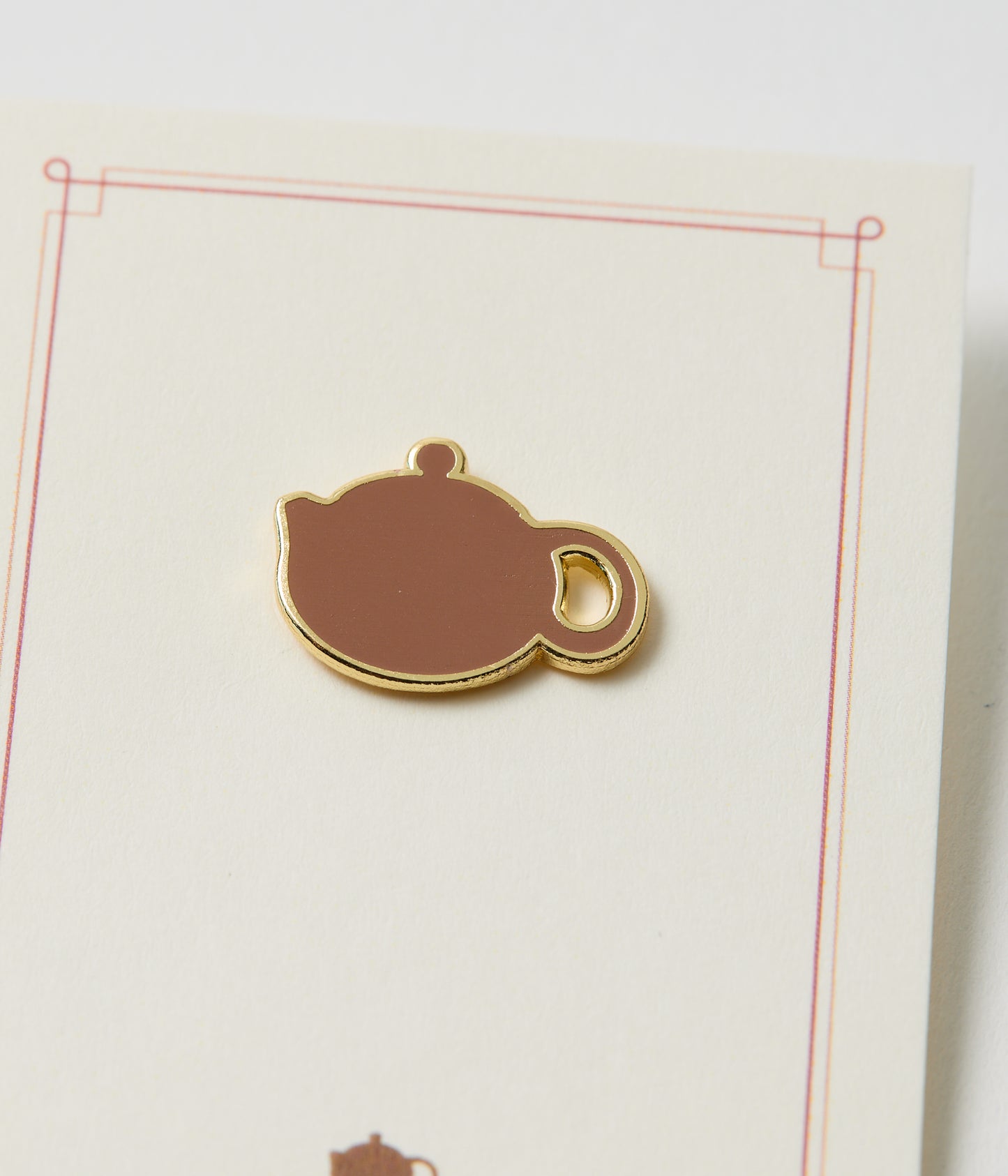 Teapot-shaped enamel pin on a white card with red border