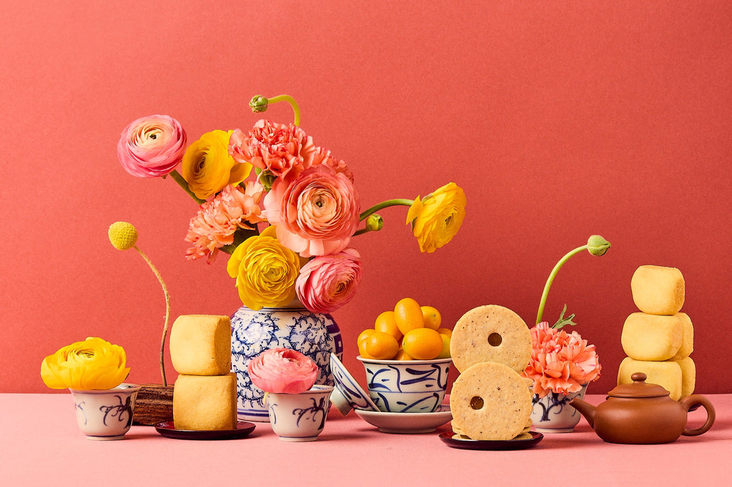 Bright floral tea table with pastries, citrus, cookies, and teapot on a coral backdrop. 