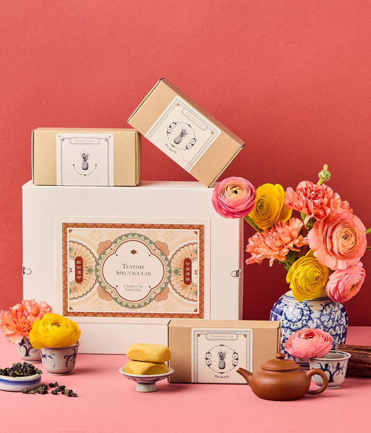 Teatime gift box with floral arrangement, tea, and pastries on a coral background.