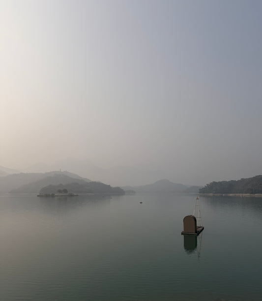 A misty lake scene with distant hills or islands, calm water, and a small floating platform with a vertical structure in the lower right.