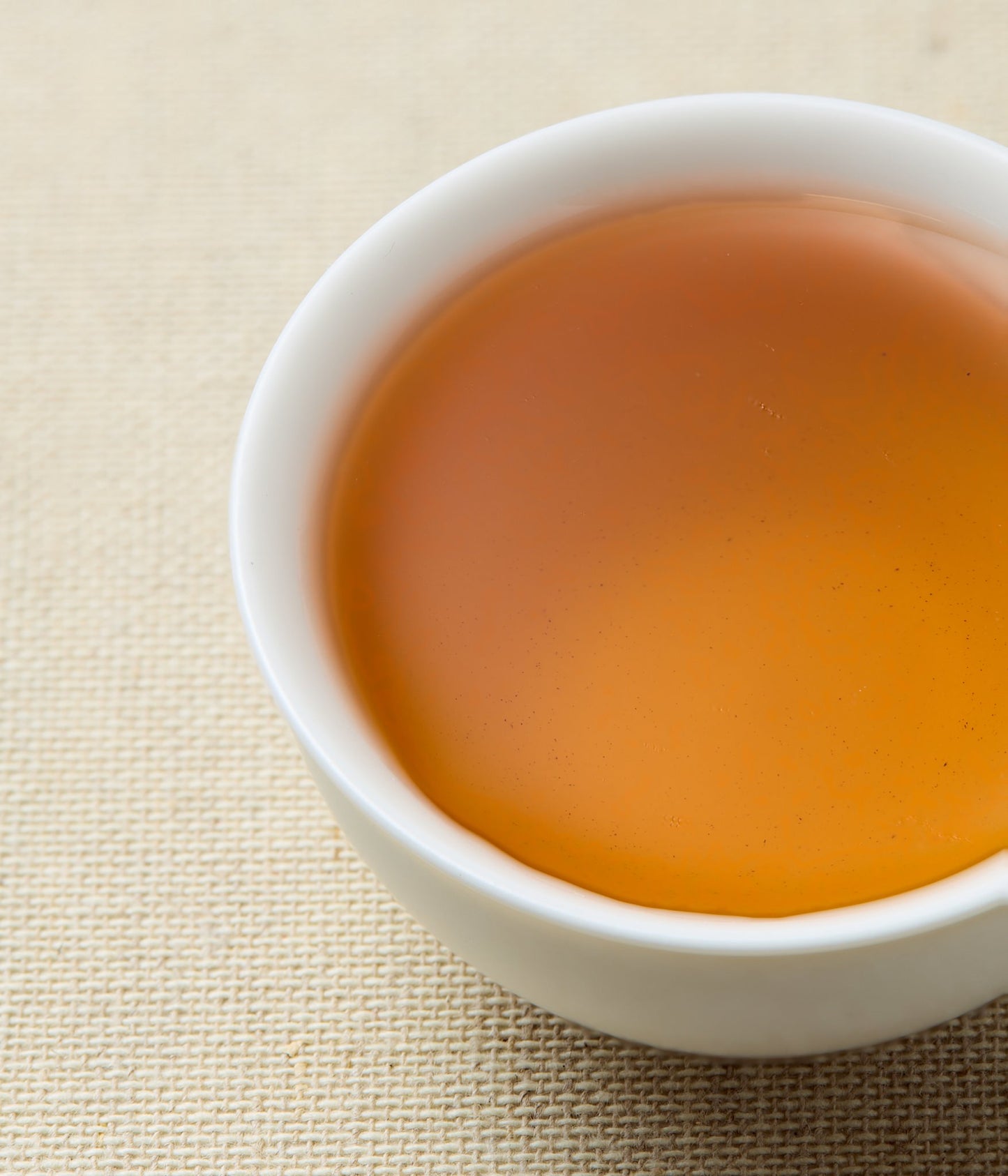 A close-up of amber-colored tea in a small white ceramic cup, set on a beige woven fabric surface.