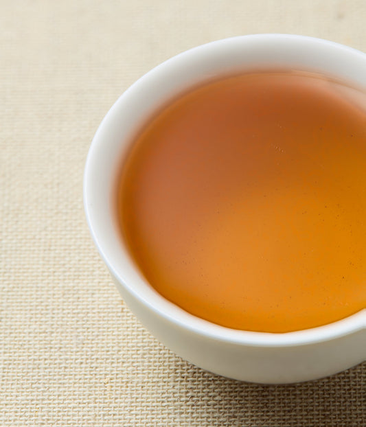 A close-up of amber-colored tea in a small white ceramic cup, set on a beige woven fabric surface.