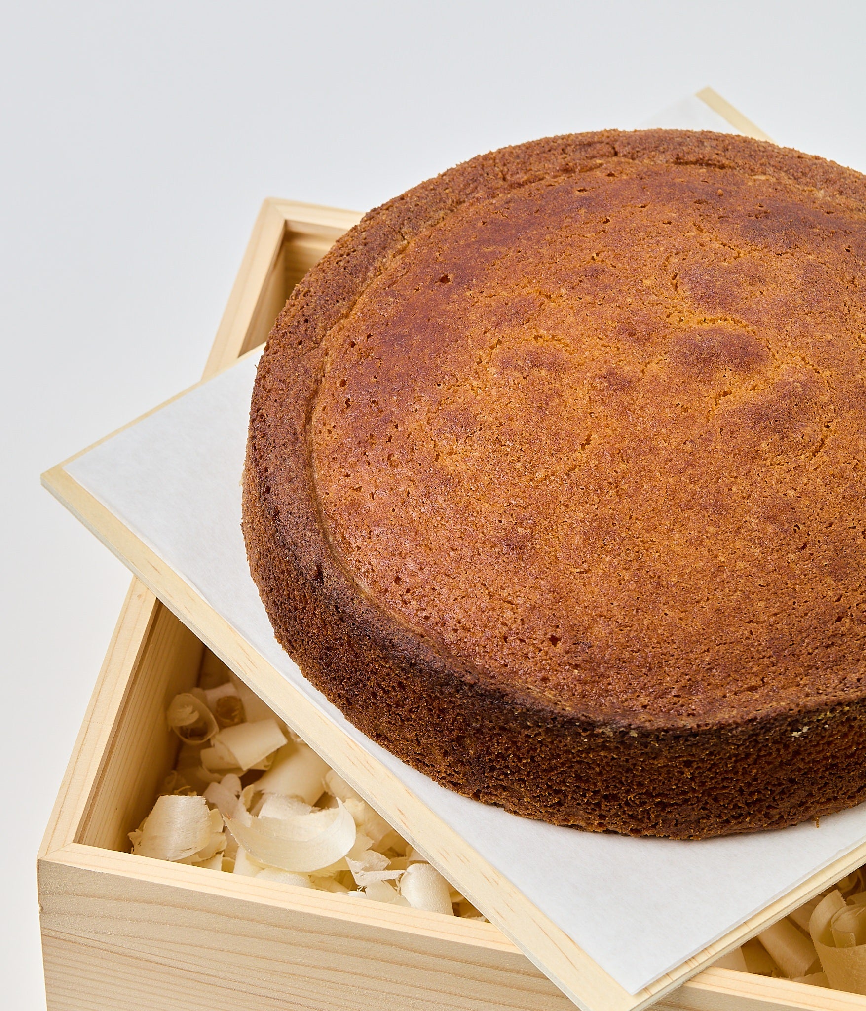 A whole round almond cake with a domed, golden-brown top, set on parchment over a light wooden board against a white background.