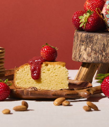 Slice of cake with strawberry sauce on a wooden plate, surrounded by strawberries and almonds.