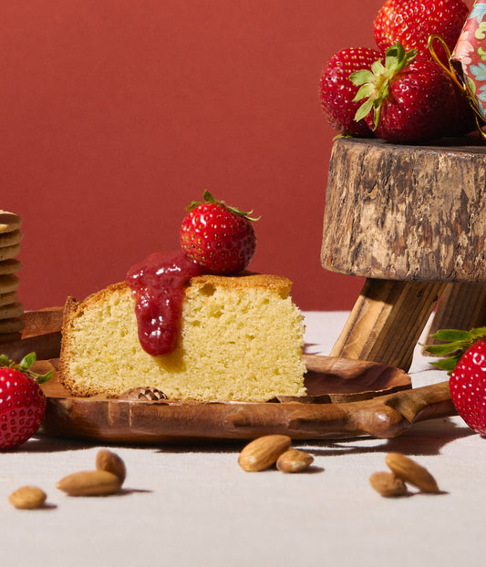 Slice of cake with strawberry sauce on a wooden plate, surrounded by strawberries and almonds.