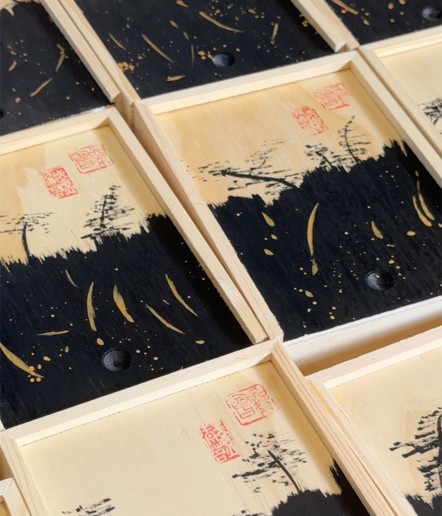 A close-up of multiple rectangular wooden boxes arranged diagonally, each featuring a black and beige landscape-style print with red seal marks and small dark circular details.