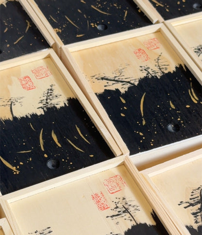 A close-up of multiple rectangular wooden boxes arranged diagonally, each featuring a black and beige landscape-style print with red seal marks and small dark circular details.