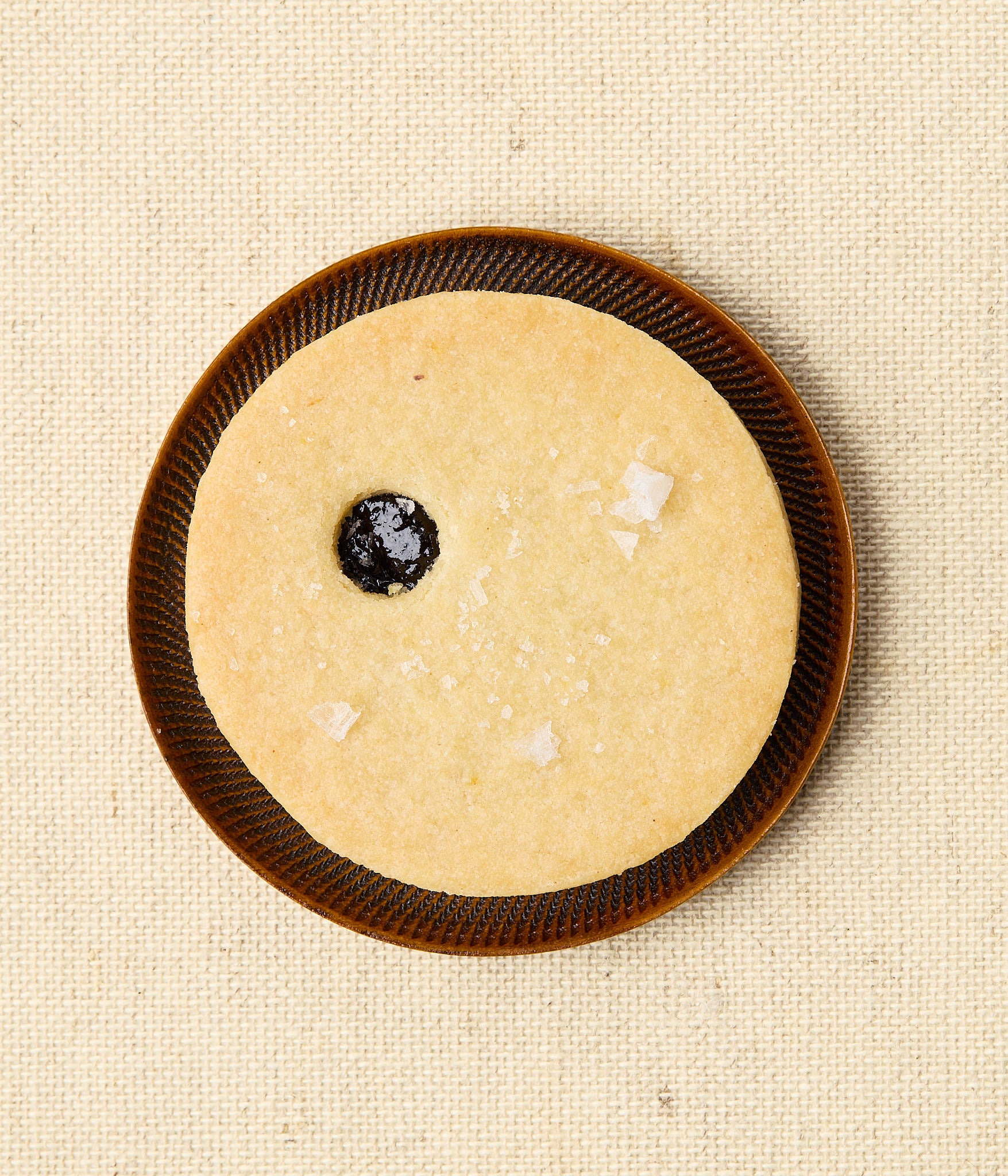 Overhead view of a round Linzer cookie on a dark ceramic plate, with a small center window revealing dark cherry jam and a scattering of flaky sea salt.