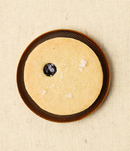 Overhead view of a round Linzer cookie on a dark ceramic plate, with a small center window revealing dark cherry jam and a scattering of flaky sea salt.