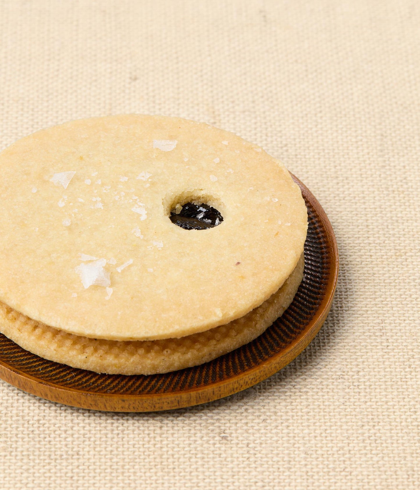 A cherry Linzer sandwich cookie on a wooden plate, the pale top cookie dusted with sea salt and a center cutout showing dark cherry filling.