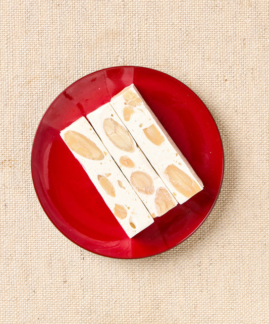 Three rectangular pieces of almond nougat with visible almond chunks arranged on a red plate, against a textured surface.