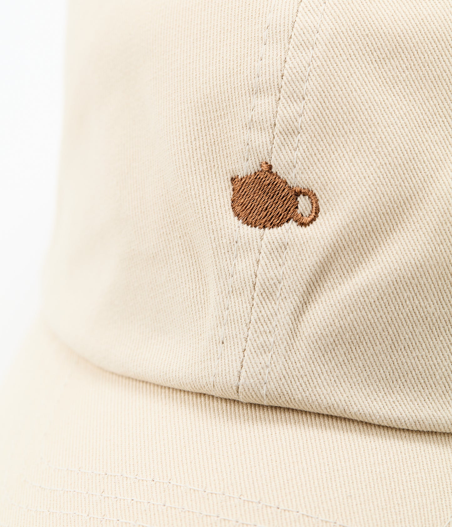 Close-up of the brown embroidered teapot logo on the front of the cream cap.