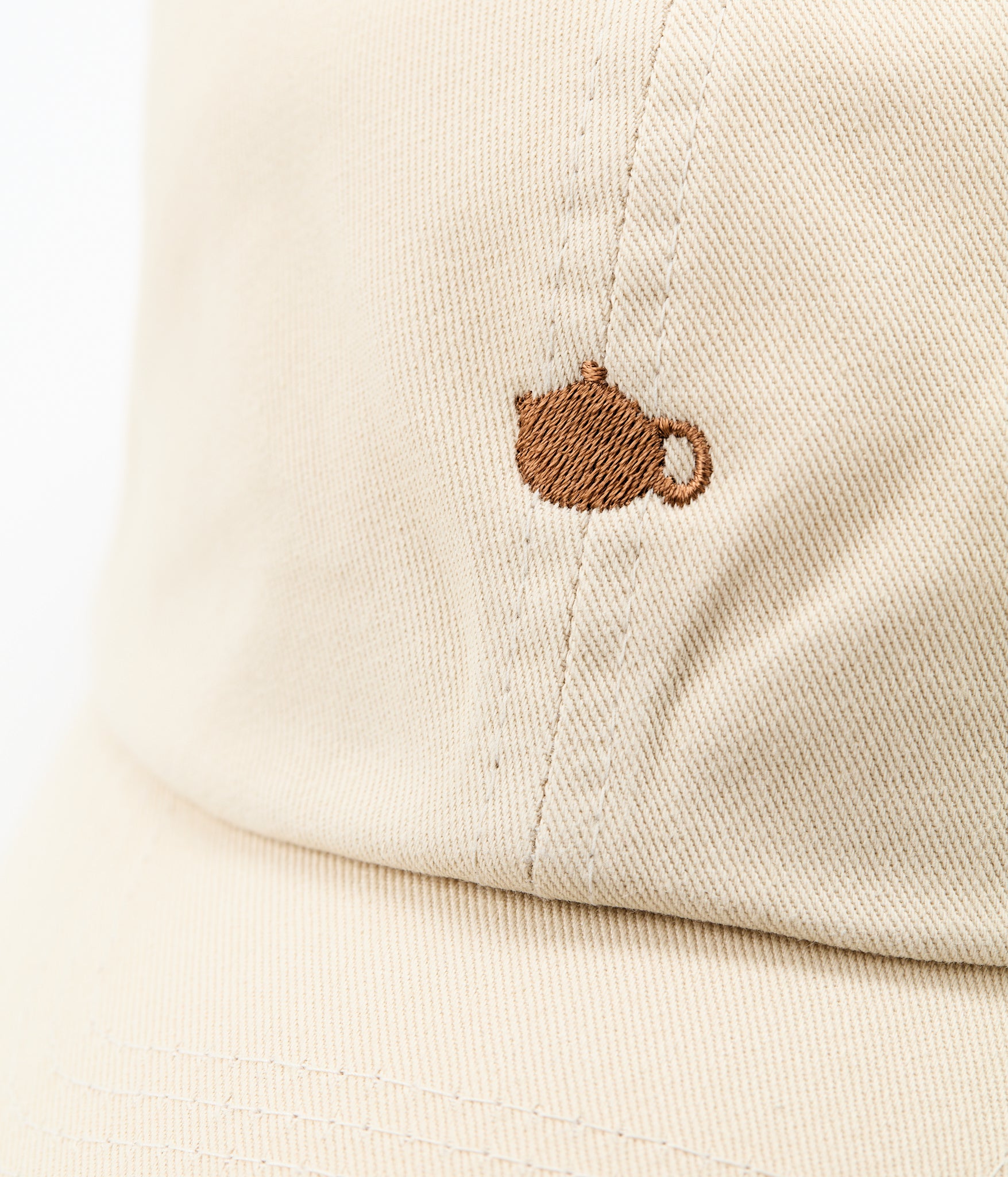 Close-up of the brown embroidered teapot logo on the front of the cream cap.