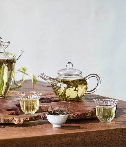 Tea-making setup with glass teapot, cups, and tea leaves on a wooden board.