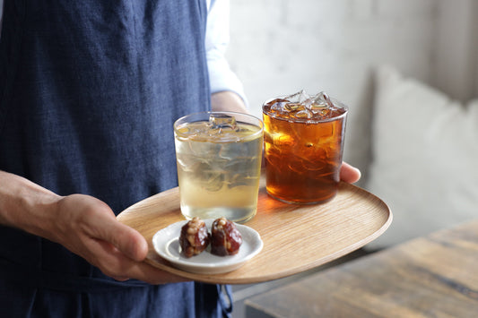 Person holding a wooden tray with two glasses of iced beverages and a small plate of snacks.