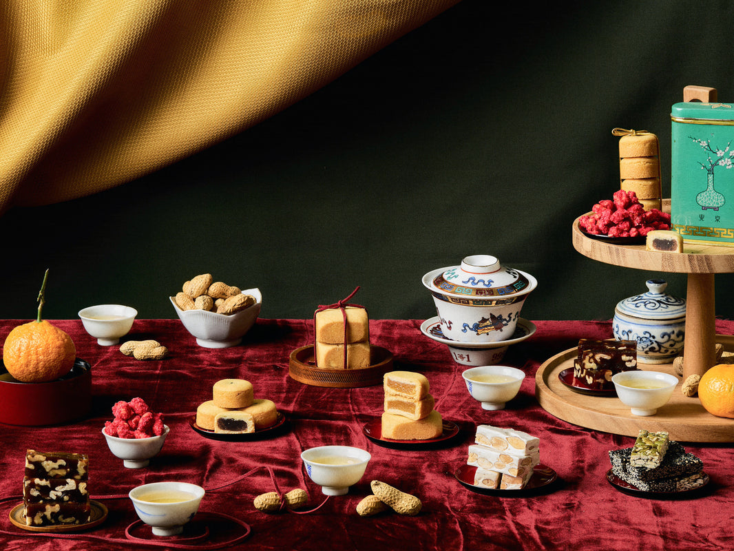 A Lunar New Year table display featuring a variety of traditional prosperity snacks, served alongside tea in porcelain cups and bowls, with vibrant red and green accents.