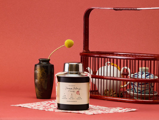 Tea canister with a decorative label on a red surface, next to a vase and a tea set.