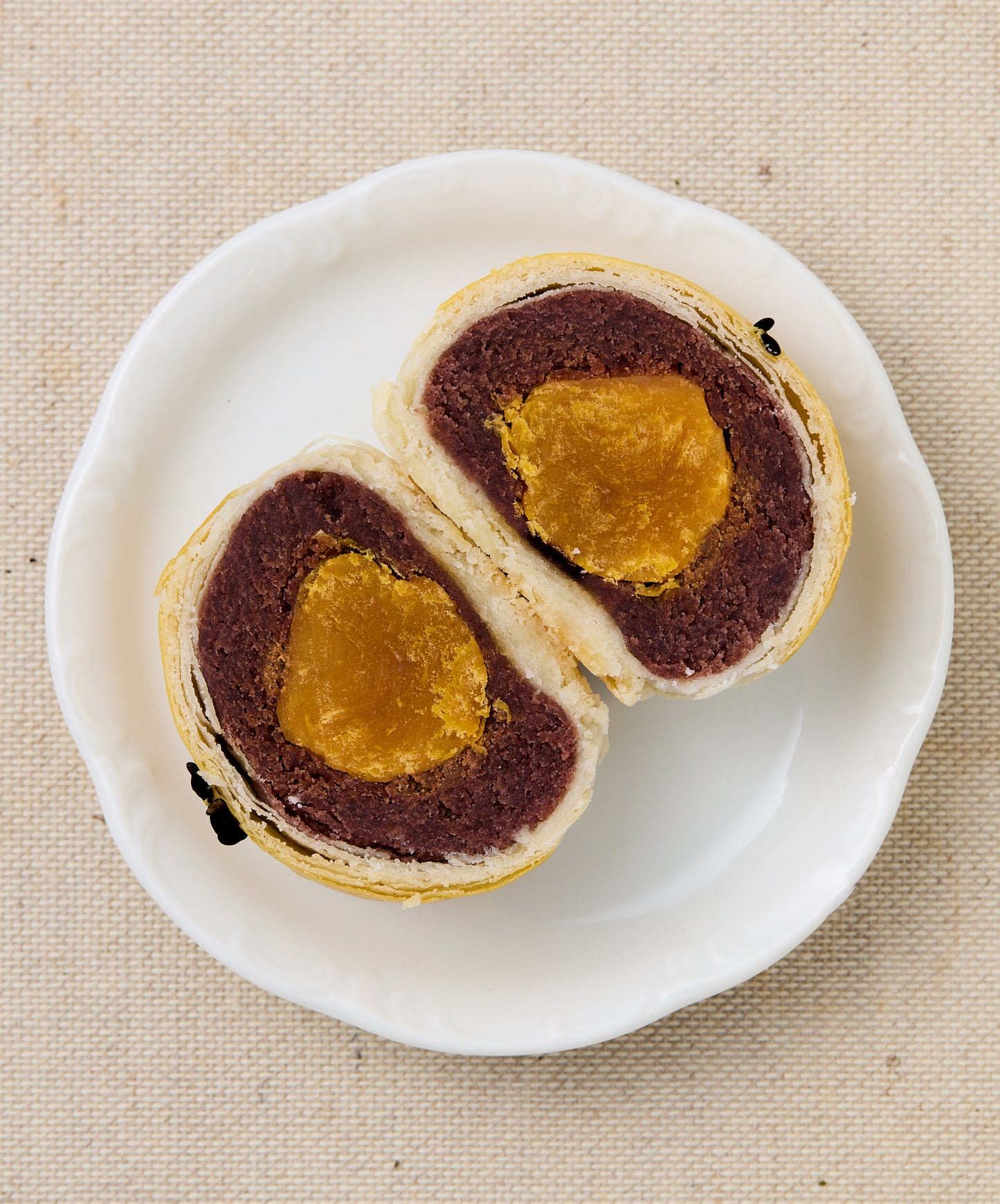 A halved mooncake revealing a dark red bean paste filling encasing a golden salted egg yolk, on a white plate.