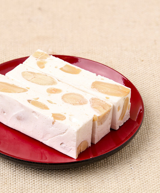 Close-up of three rectangular almond nougat bars with visible almond chunks, stacked on a red plate against a textured background.