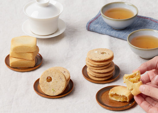 An assortment of cookies and pastries arranged on wooden plates, accompanied by two cups of tea and a white ceramic teapot on a linen tablecloth.