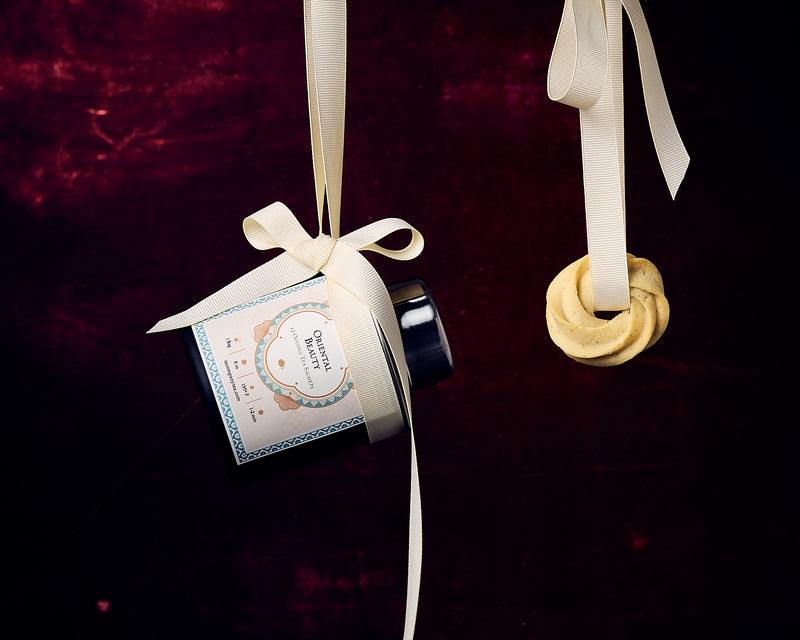 A tin labeled Oriental Beauty tied with a ribbon, suspended alongside a round cookie hanging from a matching ribbon against a dark background.