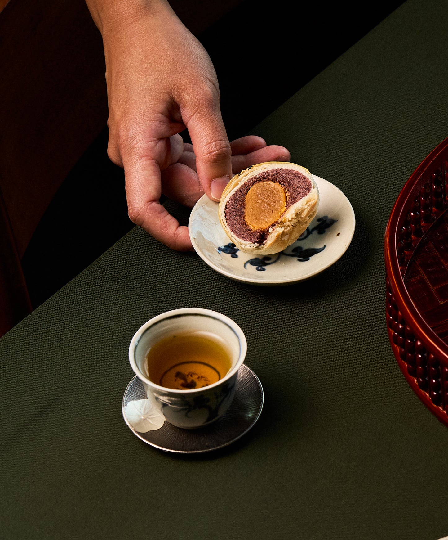 Hand holding a small plate with a mooncake next to a cup of tea on a dark surface