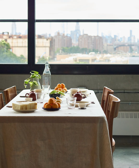 A dining table set near a large window overlooking a city skyline, adorned with fruits, teapots, and ceramic tableware.