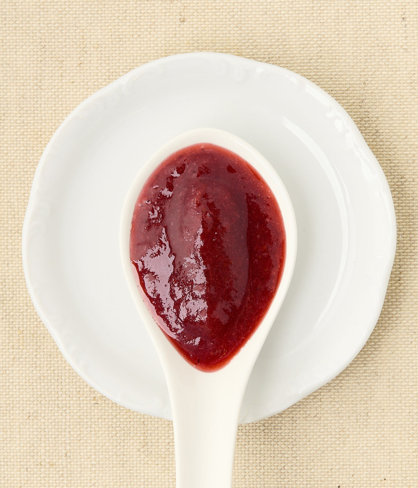 Top-down view of glossy red strawberry-rhubarb jam in a white ceramic spoon resting on a matching white dish over woven linen.