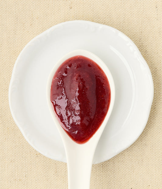 Top-down view of glossy red strawberry-rhubarb jam in a white ceramic spoon resting on a matching white dish over woven linen.