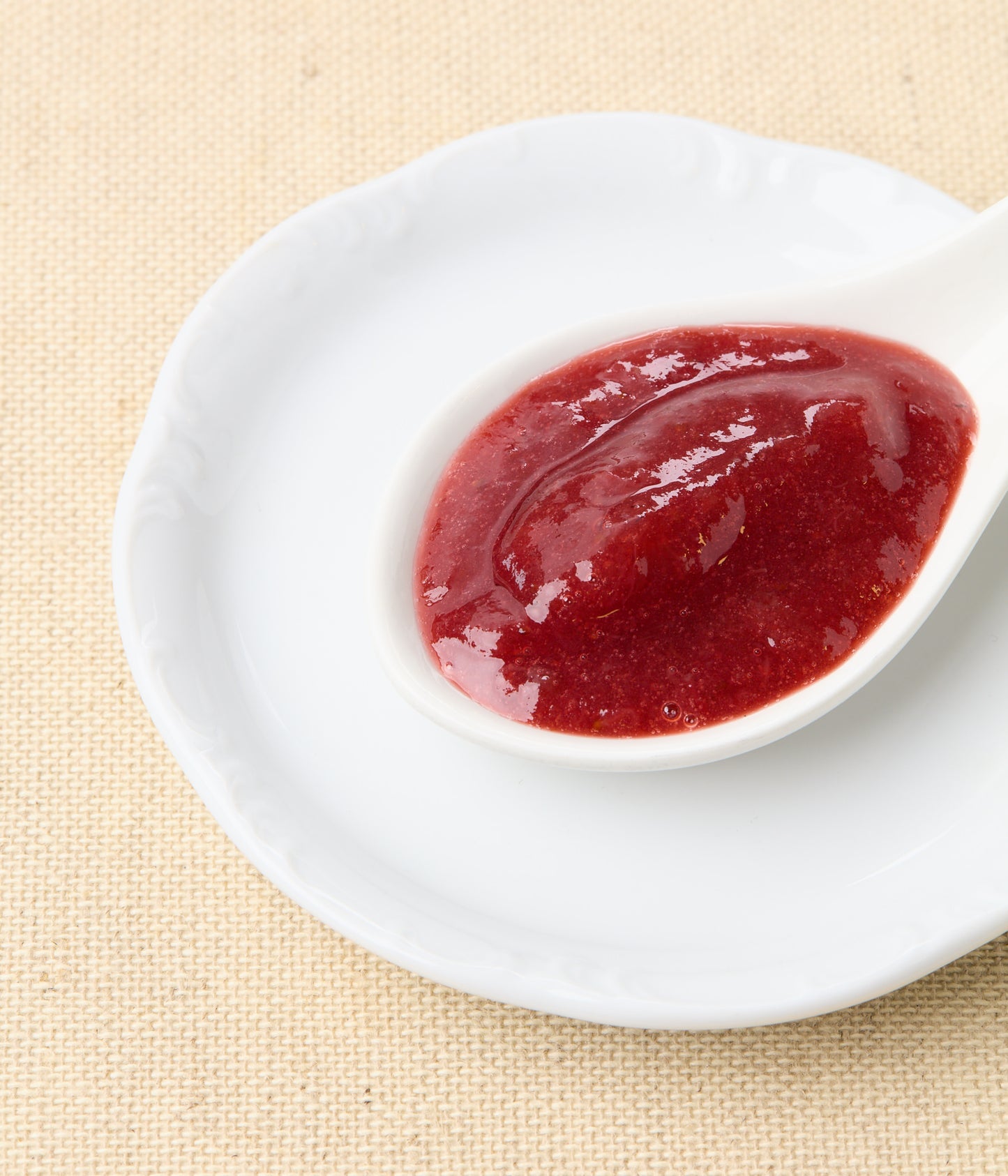 An angled close-up of bright red strawberry-rhubarb jam in a white spoon set on a white plate against a natural linen background.