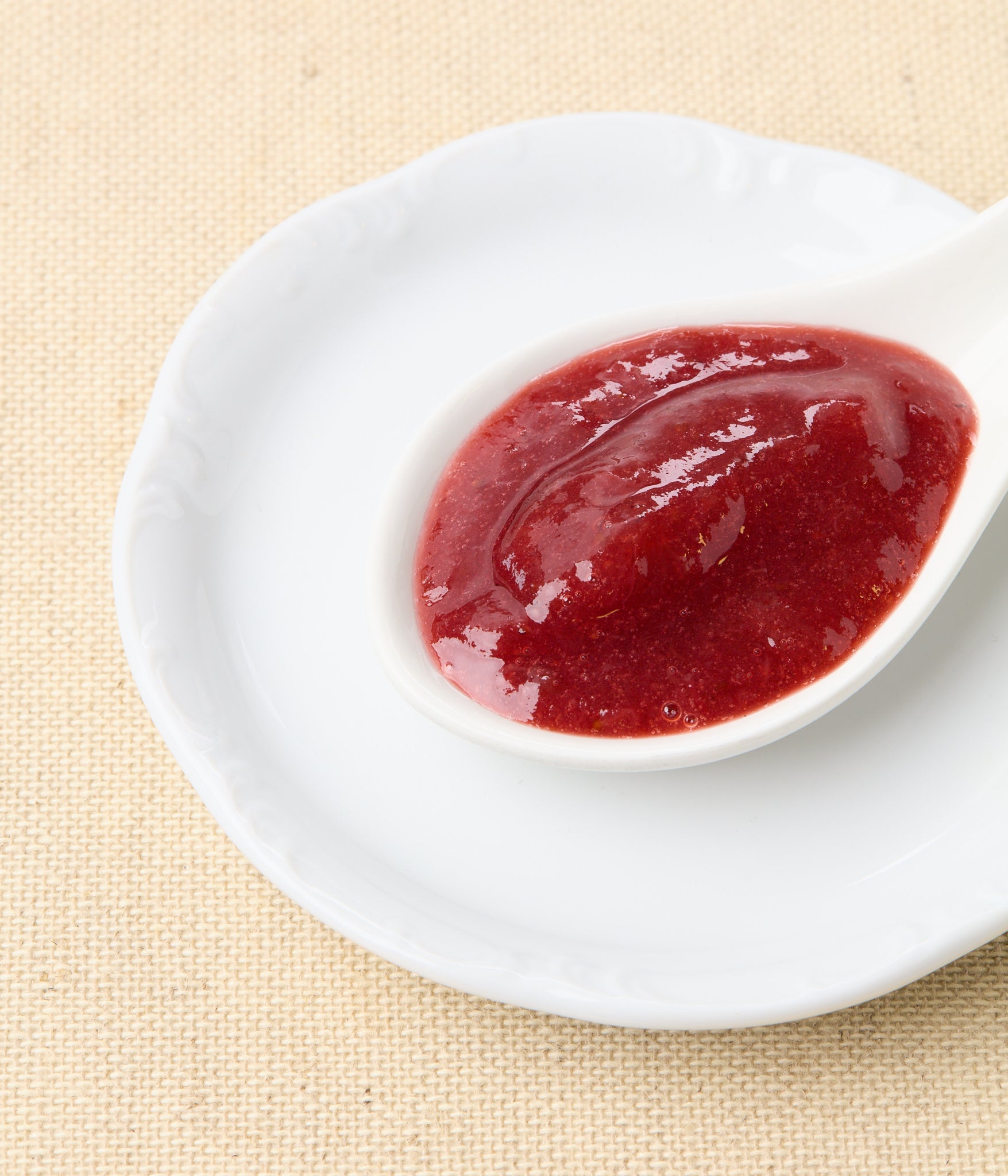 An angled close-up of bright red strawberry-rhubarb jam in a white spoon set on a white plate against a natural linen background.