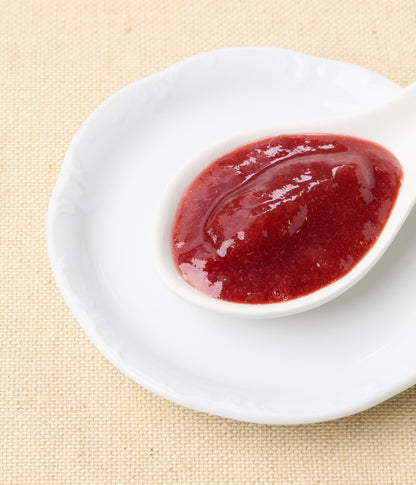 An angled close-up of bright red strawberry-rhubarb jam in a white spoon set on a white plate against a natural linen background.