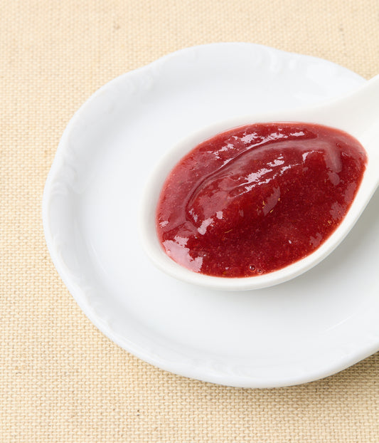 An angled close-up of bright red strawberry-rhubarb jam in a white spoon set on a white plate against a natural linen background.