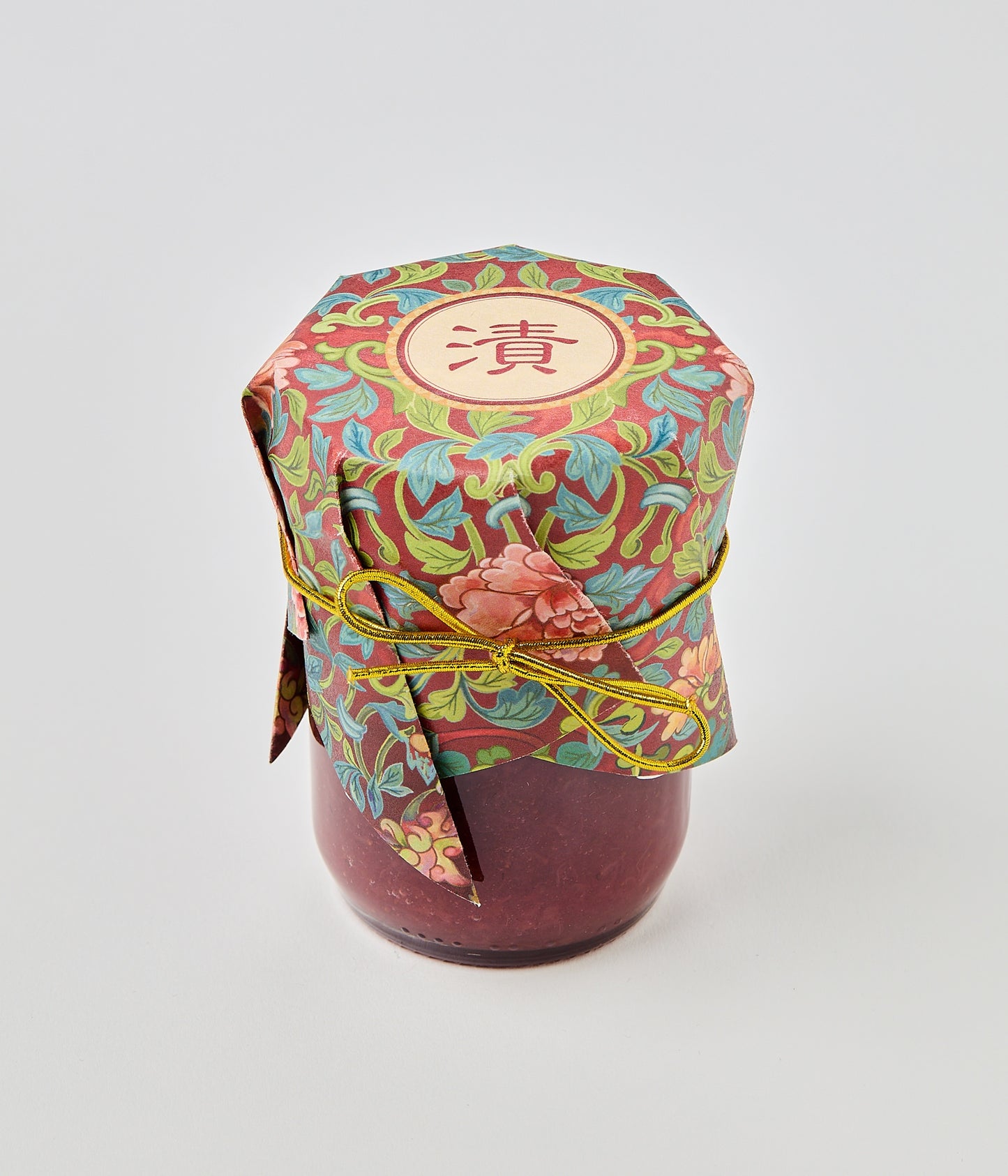 A jar of strawberry-rhubarb jam topped with floral patterned paper tied with gold cord, labeled with a red chinese character on a cream medallion.