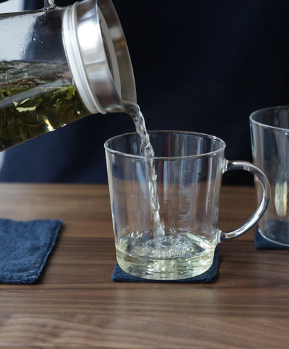 Hot tea being poured from a glass teapot into a clear glass mug on a wooden table, with blue fabric coasters and a dark background.