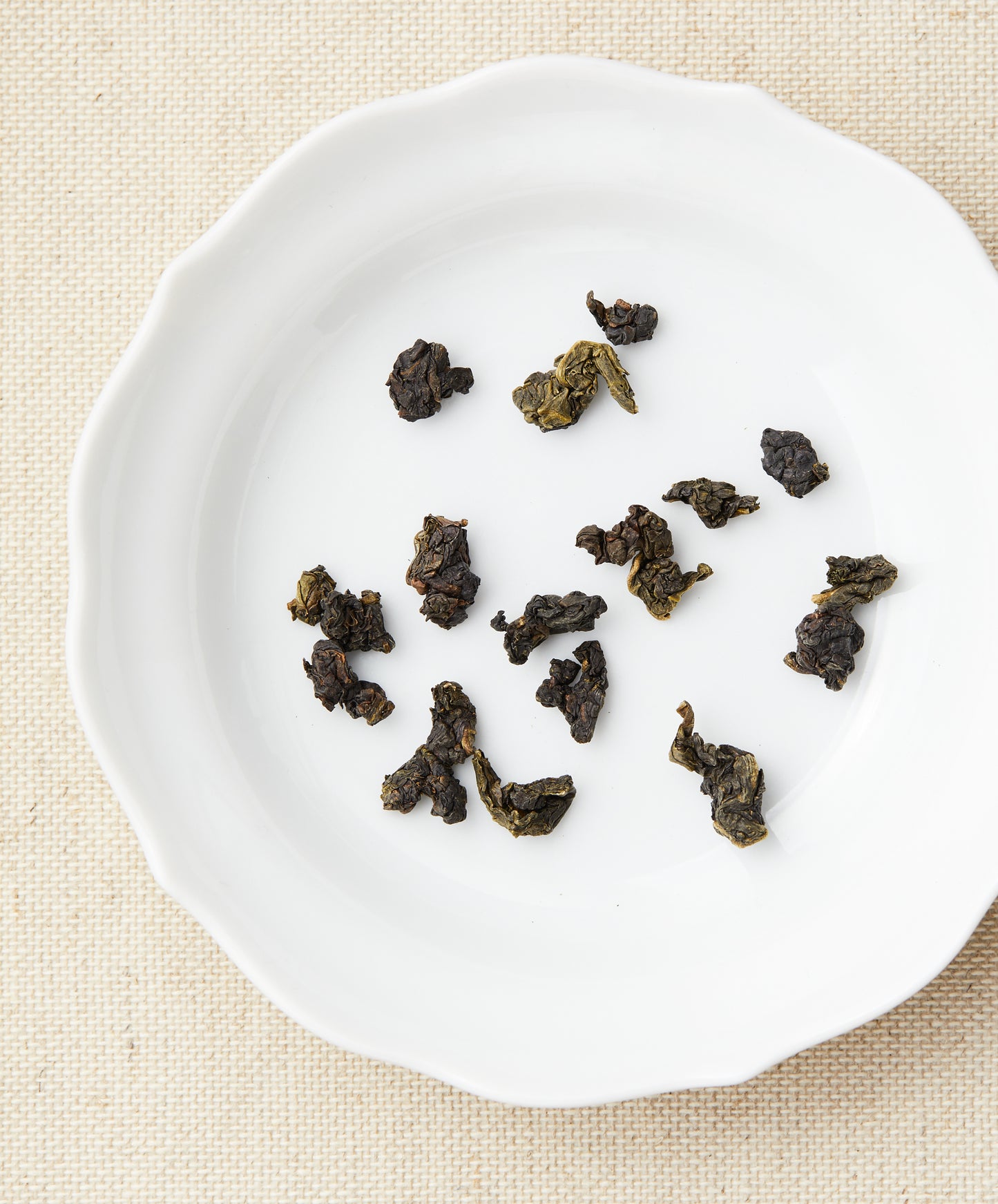 Loose-leaf tea pieces arranged on a white plate against a beige fabric background, highlighting their dark green and black hues.