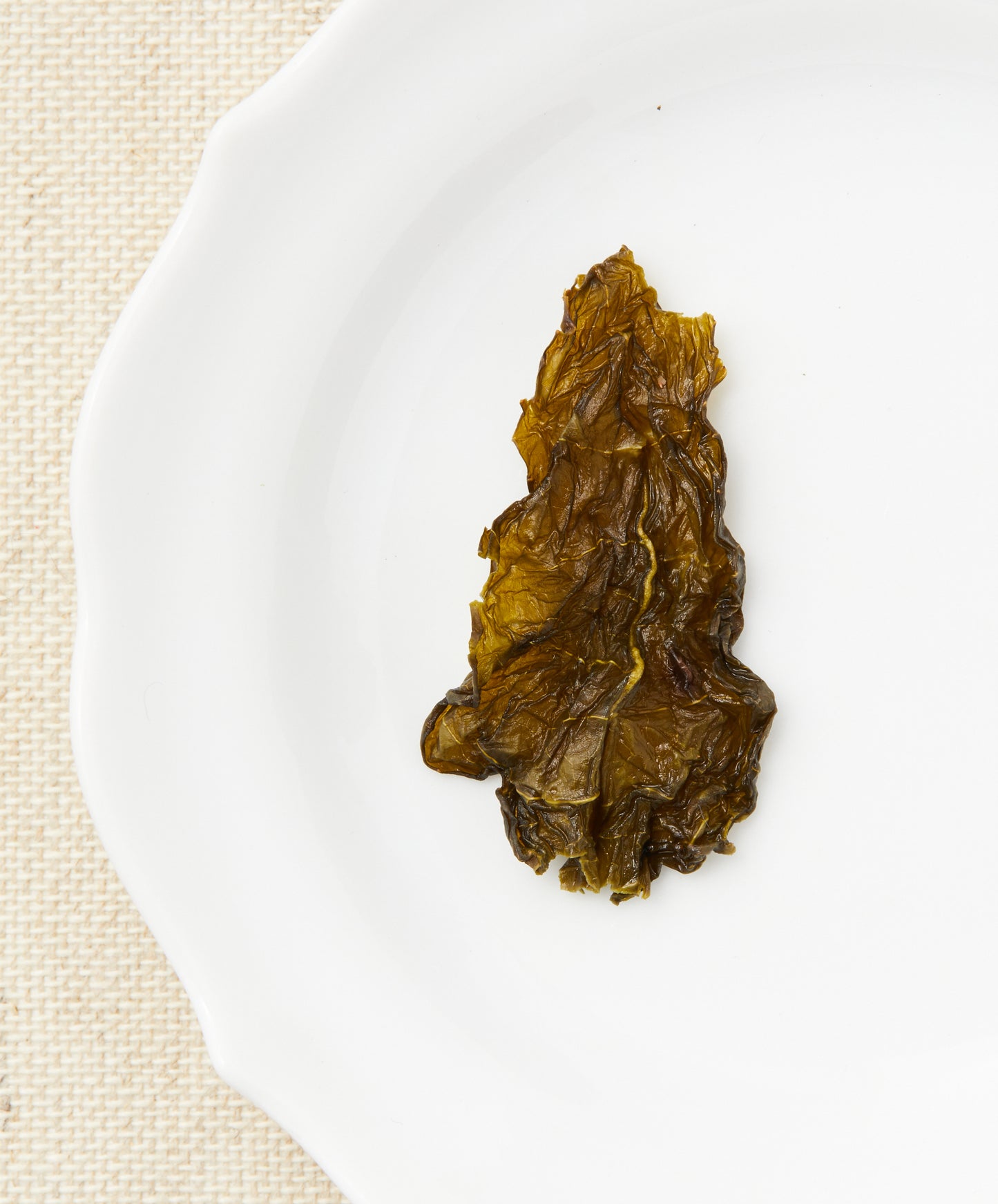 A single steeped tea leaf displayed on a white plate, showcasing its translucent texture and olive-green color.