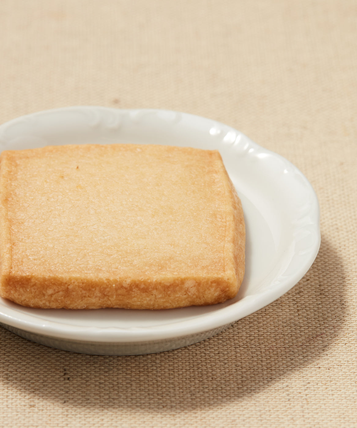 A square piece of golden shortbread placed on a white plate with a textured beige fabric background.