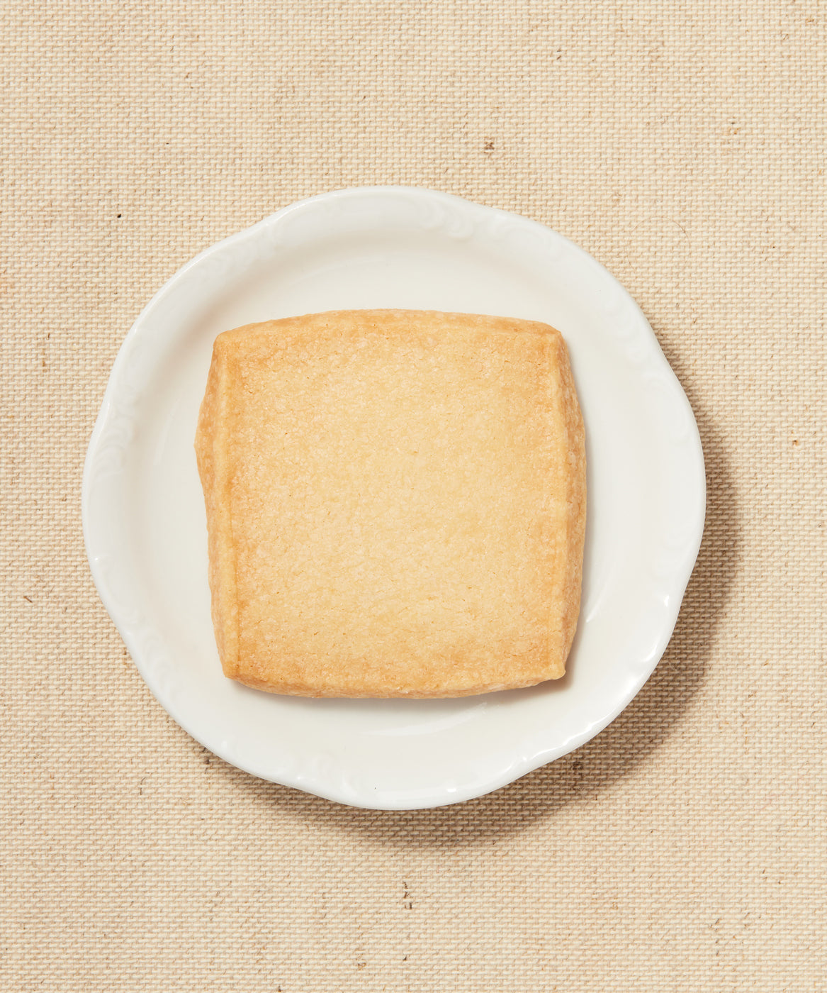 A top-down view of a square piece of shortbread on a white plate against a beige textured fabric background.