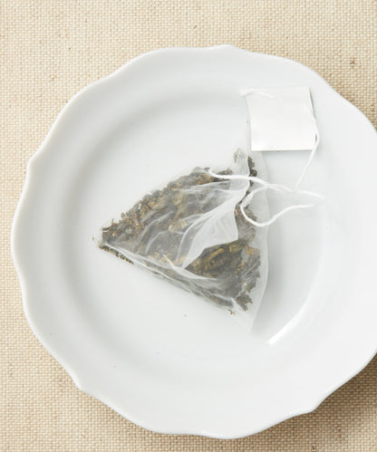 A pyramid-shaped tea sachet filled with dried leaves, displayed on a white plate against a beige background.