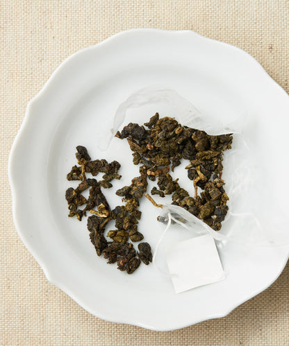 A pyramid-shaped tea sachet with loose leaves scattered around it on a white plate.