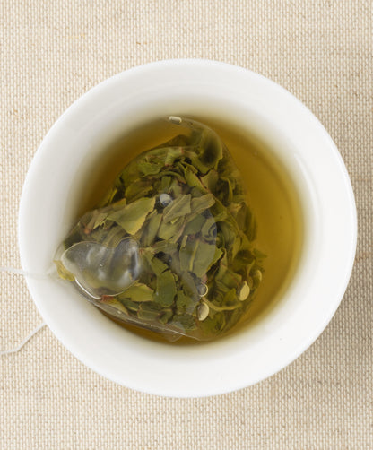 A cup of steeped tea with a pyramid-shaped tea sachet containing loose leaves, submerged in the liquid, set against a beige fabric background.